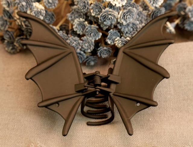 A brown metal hair clip designed to resemble bat wings, displayed against a floral background.