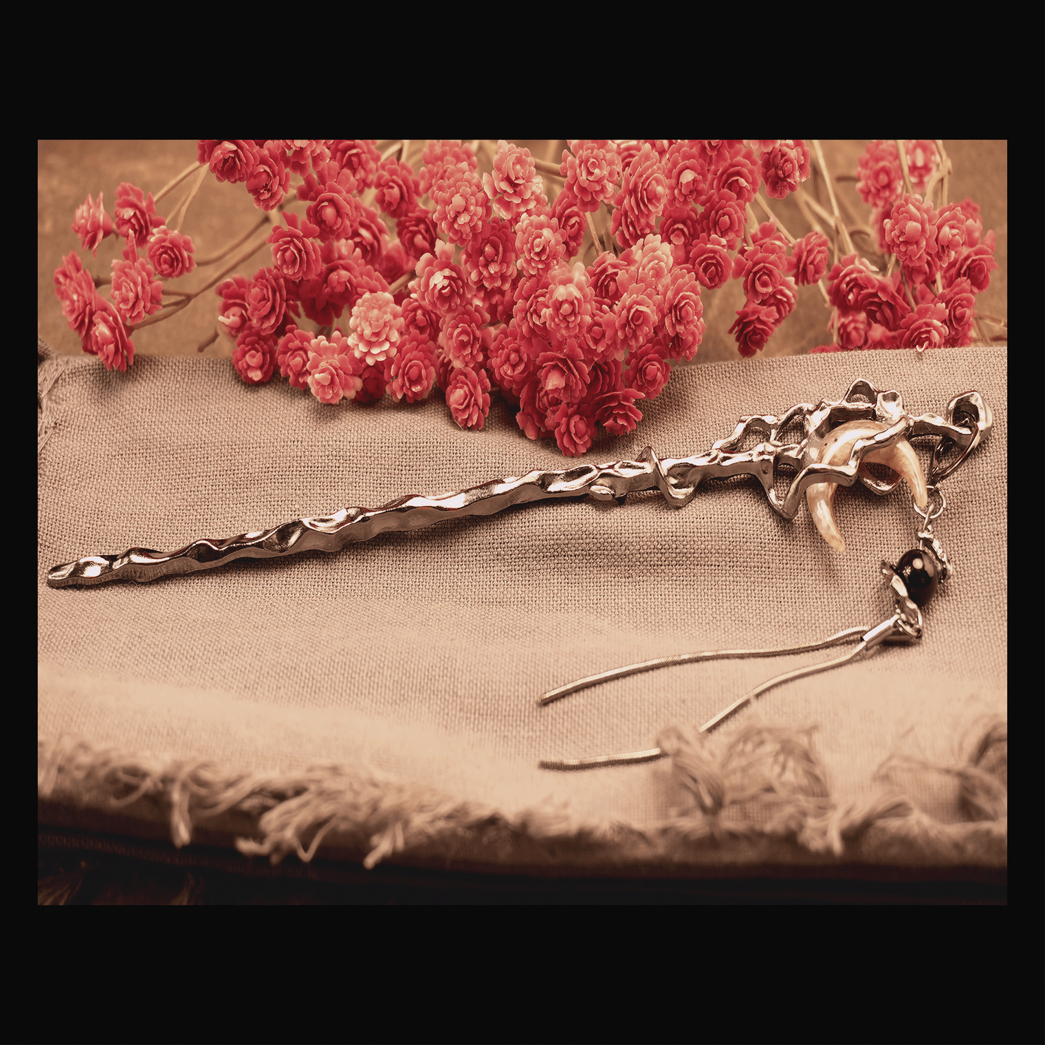 A silver-colored hair pin shaped like a crescent moon with a tassel, displayed on a beige fabric surface with red flowers in the background.