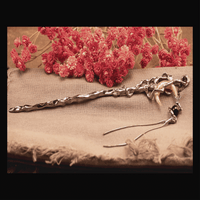 A silver-colored hair pin shaped like a crescent moon with a tassel, displayed on a beige fabric surface with red flowers in the background.