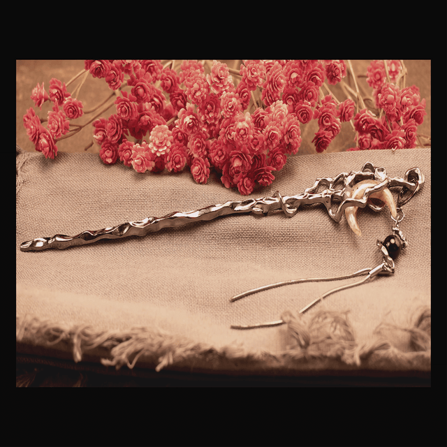 A silver-colored hair pin shaped like a crescent moon with a tassel, displayed on a beige fabric surface with red flowers in the background.