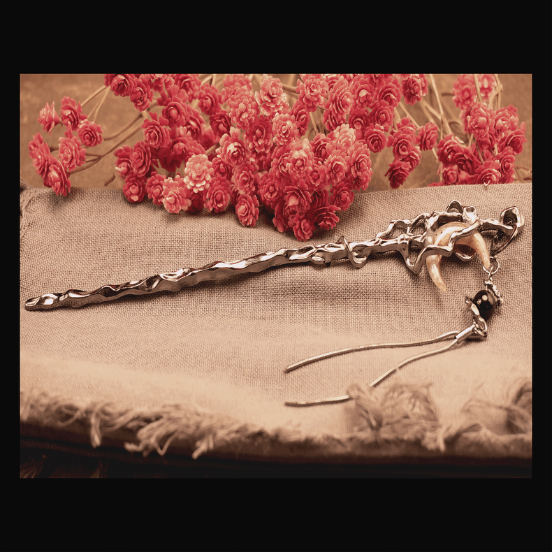 A silver-colored hair pin shaped like a crescent moon with a tassel, displayed on a beige fabric surface with red flowers in the background.