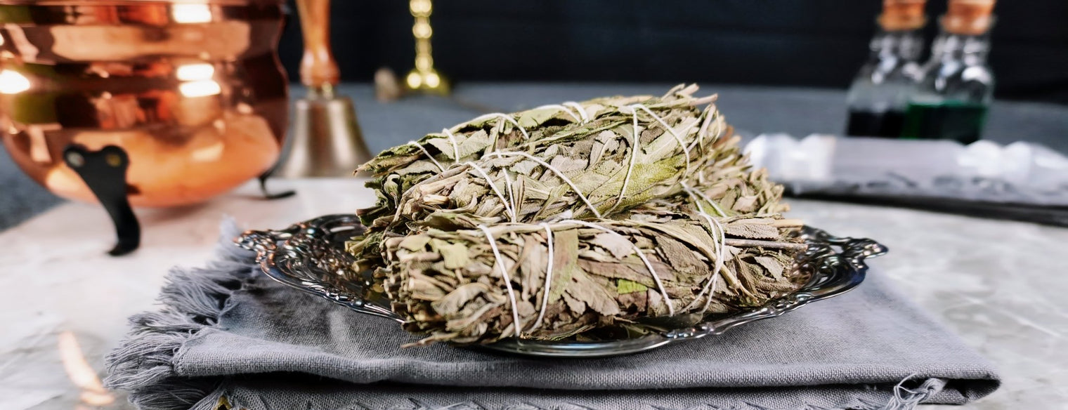 A bundle of mugwort smudge sticks placed on a dish with a smoky appearance, set on a table with various items in the background, including a copper container.