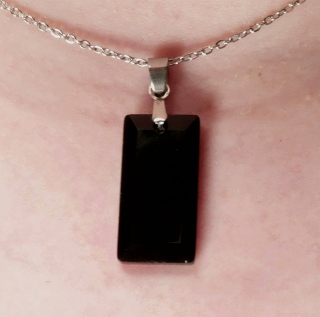 A black faceted obsidian crystal pendant necklace suspended from a silver chain, displayed against a partial view of a person's neck.
