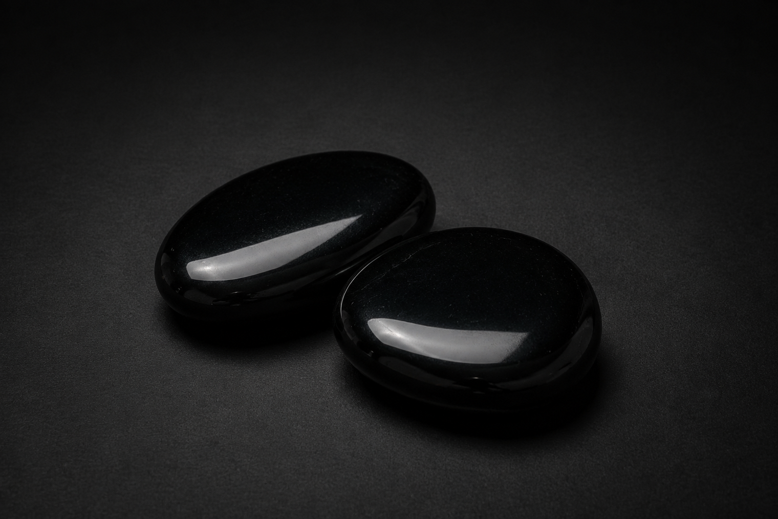 Two polished jet-black Obsidian palm stones with glossy reflections on a matte black surface