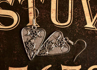 A pair of silver-colored pendant earrings featuring a planchette design with a crescent moon and a crow, displayed against a dark background with ornate patterns.