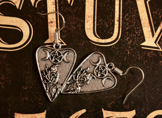 A pair of silver-colored pendant earrings featuring a planchette design with a crescent moon and a crow, displayed against a dark background with ornate patterns.