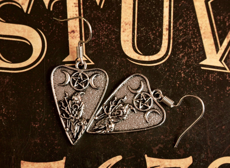 A pair of silver-colored pendant earrings featuring a planchette design with a crescent moon and a crow, displayed against a dark background with ornate patterns.