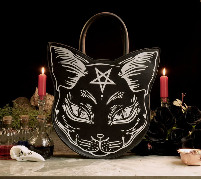 A black handbag in the shape of a cat with a pentagram on its forehead, presented in a mystical and gothic style setting with candles and vintage decorations.