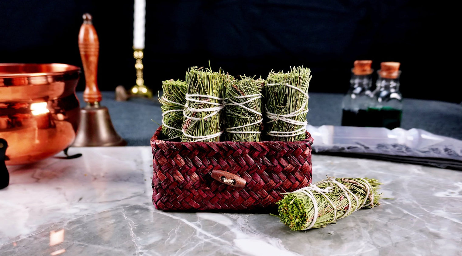 A bundle of pine herb wands tied with string, displayed in a woven basket on a table with various items in the background.