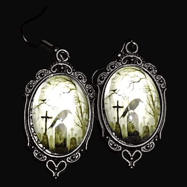 A pair of black oval earrings with a raven perched on a tombstone design, set against a white oval frame with intricate detailing.