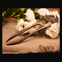 A brown metal hair pin with a raven design, displayed on a beige fabric surface with white flowers in the background.