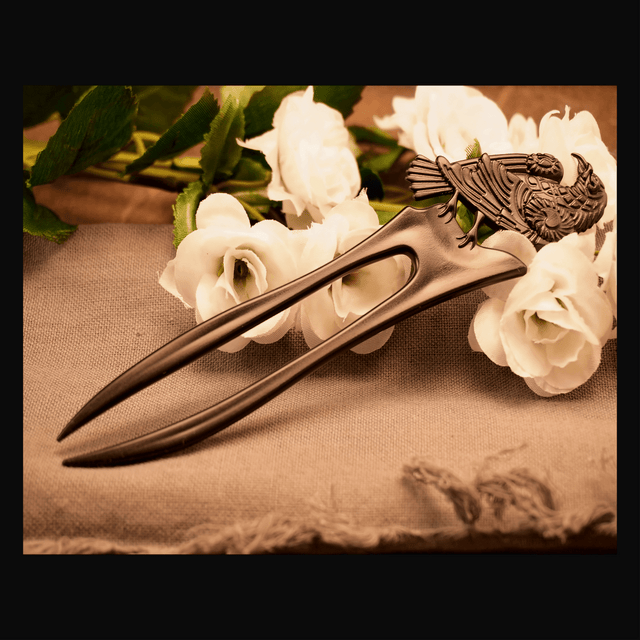 A brown metal hair pin with a raven design, displayed on a beige fabric surface with white flowers in the background.
