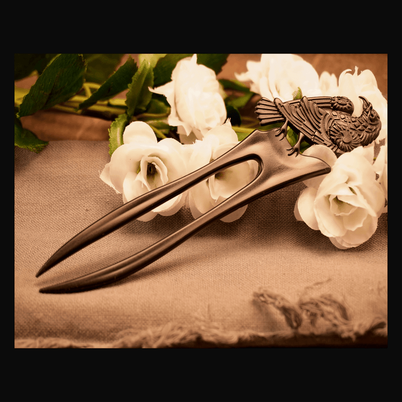 A brown metal hair pin with a raven design, displayed on a beige fabric surface with white flowers in the background.