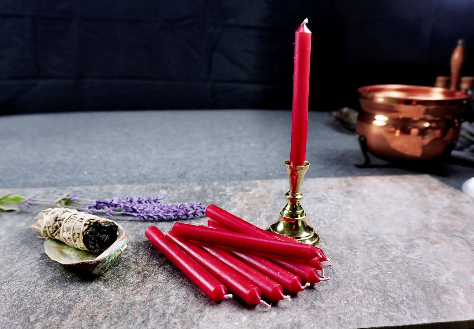 A group of red chime candles arranged on a gray surface with a lighted candle on a gold holder in the background, and a small white and green object to the left.