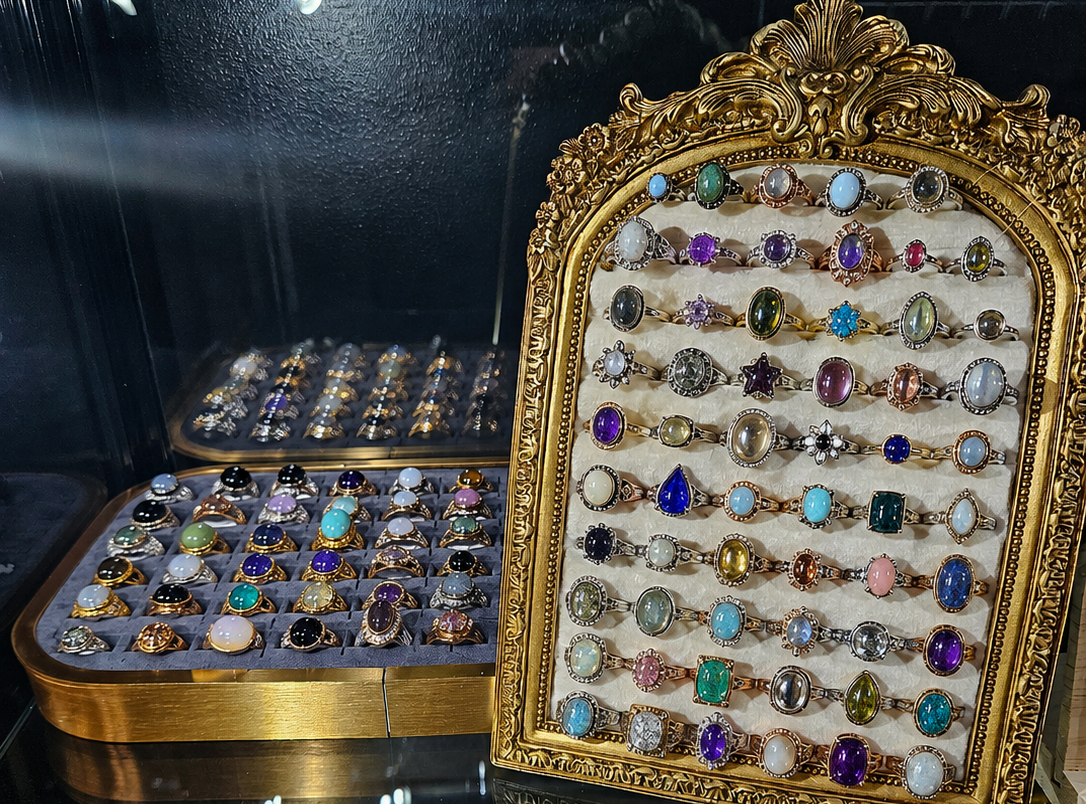 A display case filled with assorted rings, crystals, and small jewelry items. A large ornate gold-framed board on the right holds many colorful rings, with additional trays of rings and stones visible behind and below it.