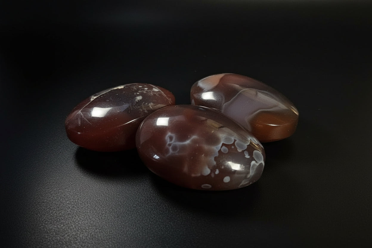 Three polished agate stones with natural banding in reddish-brown, orange, and white, displayed on a dark surface against a dark gray background.