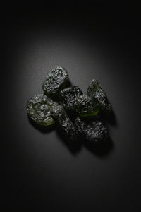A cluster of small, dark green to black, semi-translucent, rock-like pieces with a rough, speckled texture, resting on a white sheet of paper over a dark wood surface.