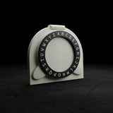 A white plastic alphabet ring holder with a black circular ring printed with uppercase alphabet letters, resting on a dark brown wooden table.
