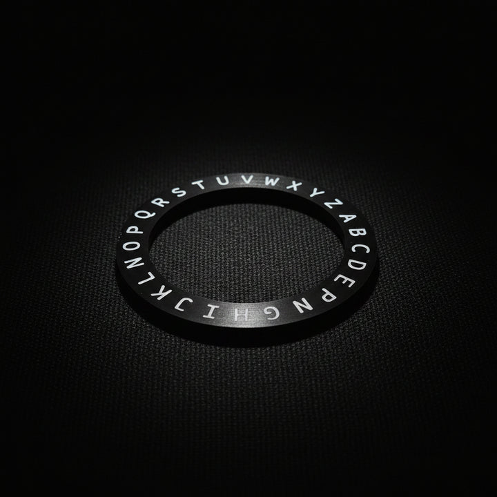A black circular ring with white uppercase letters arranged in a circle, placed on a dark brown wooden surface.