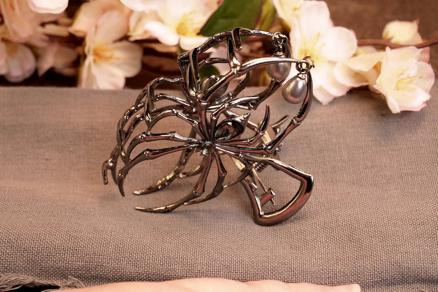 A metal hair clip with a intricate, dark, spider-like design, displayed on a fabric surface with floral patterns in the background.