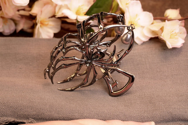 A metal hair clip with a intricate, dark, spider-like design, displayed on a fabric surface with floral patterns in the background.