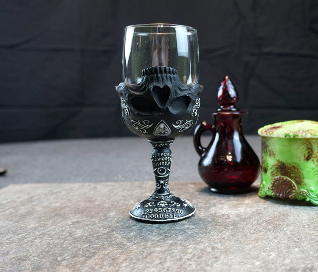A black ceramic goblet with a spirit board and OCCULT script design, displayed on a table with a small red bottle and a green object in the background.