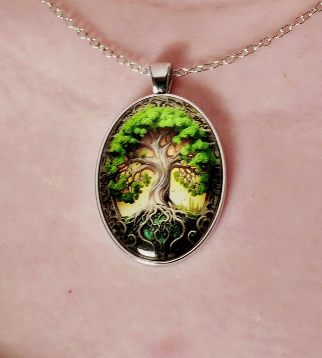 An oval-shaped pendant necklace with a detailed illustration of the Tree of Life design, featuring green leaves and brown branches on a black background.