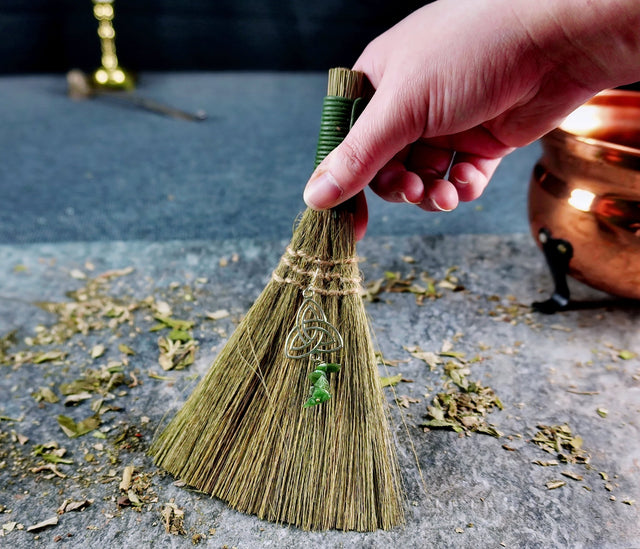 A hand holding a small, hand-bound broom with a Triquetra charm attached to the handle, set against a background with scattered leaves and a copper container.