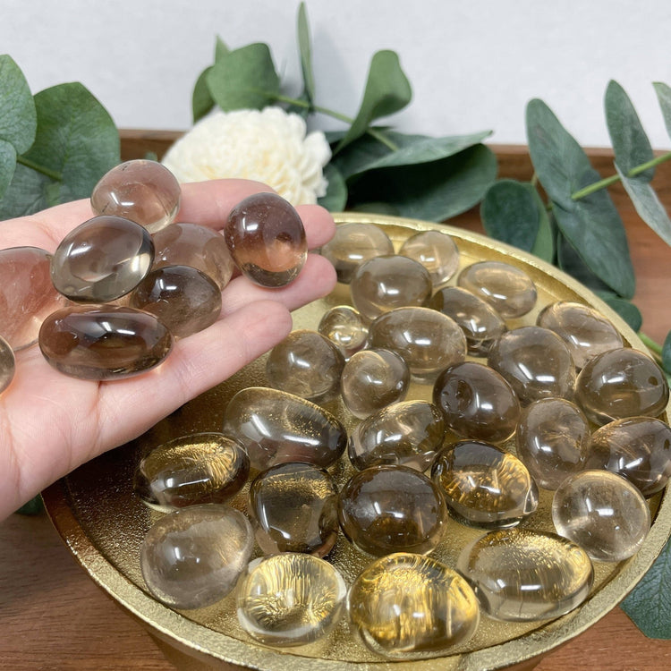 Hand holding a clear crystal rock over a gold dish filled with similar crystals, with greenery in the background.