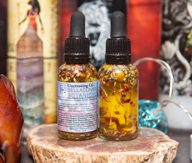 Two bottles of 'Uncrossing Oil' by Belladonna Botanica on a wooden surface with a colorful background.