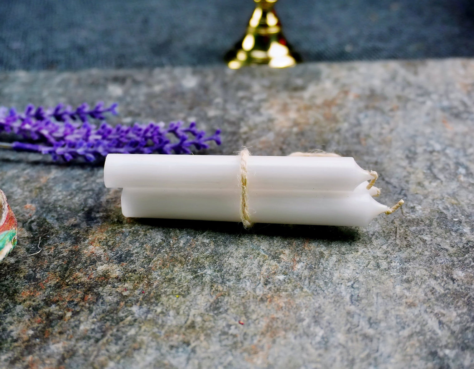 A white ritual chime candle placed on a stone surface, tied with a string, surrounded by purple flowers and a gold-colored bell in the background.