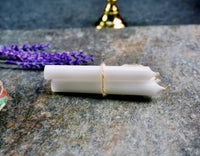 A white ritual chime candle placed on a stone surface, tied with a string, surrounded by purple flowers and a gold-colored bell in the background.